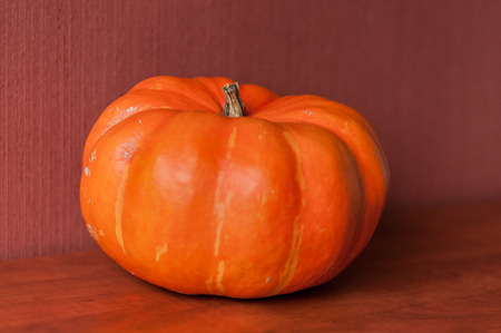 An orange pumkin is lying on a brown kitchen table, isolated on a red brown background.