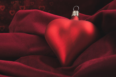 A red christmas bauble in the shape of a heart lies on a soft velvet blanket and shiny little hearts in the background.の写真素材