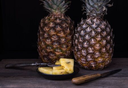 Sliced pineapple on old rustic desk with knife and with whole pineapple behind. Black background.の写真素材