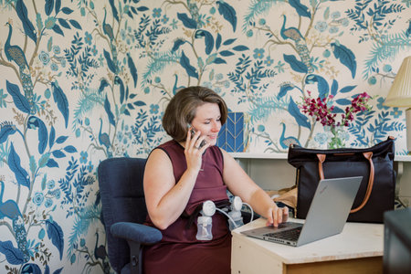 Caucasian working mother talks on phone at desk while pumping.の写真素材