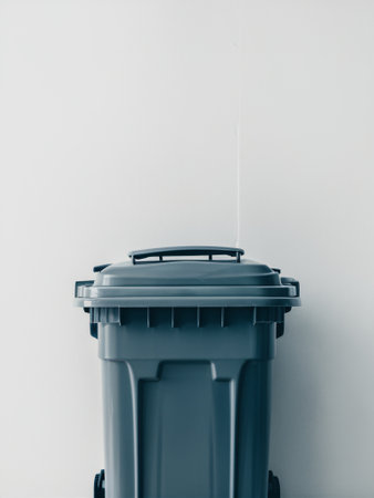 A modern gray trash bin positioned against a clean white wall, emphasizing cleanliness and organization.の素材