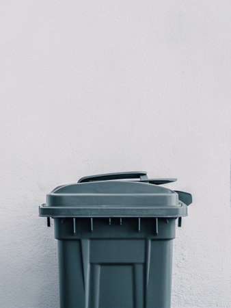 A dark gray trash bin against a plain white wall, symbolizing waste management and cleanliness.の素材