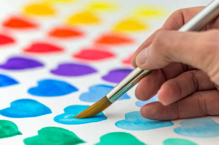 Multicolored hearts being painted with a watercolor paintbrush and paint with hand holding the brush. Shallow depth of field.の写真素材