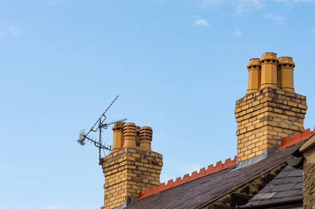 Old residential rooftop and chimneys with TV ariel. Slate roof, decorative red terracotta ridge tiles, yellow brick chimney stacks and chimney pots. Blue sky background with copy space.の写真素材