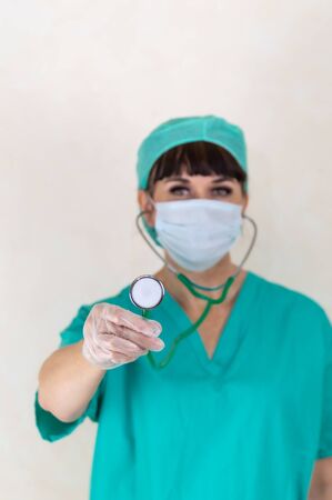 Blurred female doctor or nurse wearing face mask, hair net and green scrubs. Focus on stethoscope and hand with latex glove. Making eye contact. Plain pale background with copy space.の写真素材