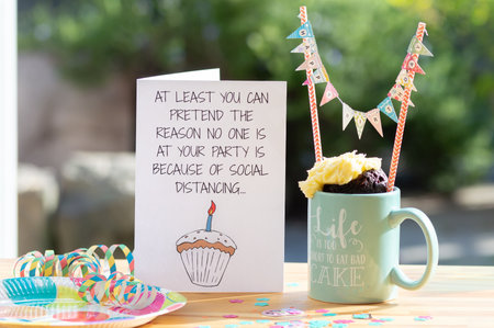Wrexham, UK - May 6, 2020: Birthday celebration for someone isolating alone during the coronavirus pandemic lockdown. Funny card with text about social distancing, and mug cake for one.