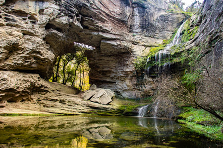 A scenic view of a rocky cave with a waterfall and a calm pond.