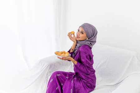 A young Malay lady with hijab head covering, celebrates end of Ramadan, dressed in purple kebaya dress, holding traditional bahulu cake, sitting on white sofa, white background, waist up portraitの写真素材