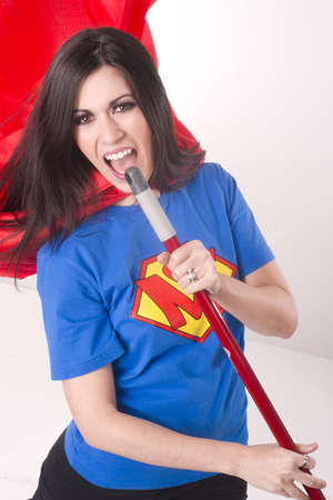 A beautiful woman in crested shirt and red cape lipsyncs with her broom stick while cleaningの写真素材