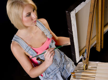 A Happy Blond Woman Dressed in Overalls Provides Finishing Touches to Painting on Easelの写真素材