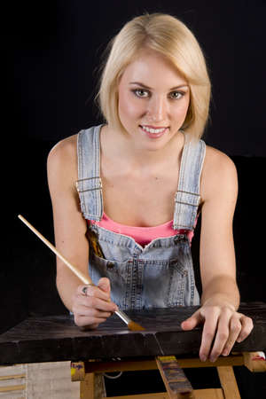 A Happy Blond Woman Dressed in Overalls Provides Finishing Touches to Painting on Easelの写真素材