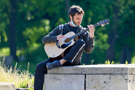Young man playing guitar sitting on stone planks in the park