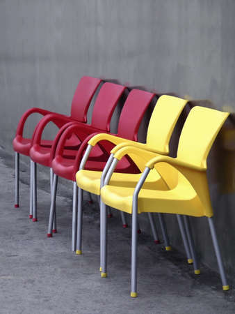 Five plastic chairs, three red and two yellow, aligned in a row next to a concrete gray wall.の写真素材