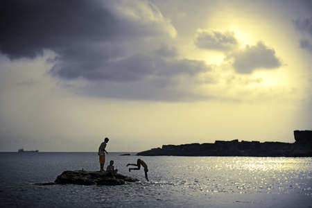 Peaceful beach, cloudy sky and silhouettes of kids divingの写真素材