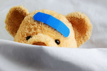 Photo of a sick teddy bear with a blue bandage in bedの写真素材