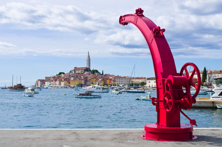 Explore the stunning Croatian coast. A vibrant port with clear blue water, boats, and a vintage red crane against the backdrop of a charming historic town.の写真素材