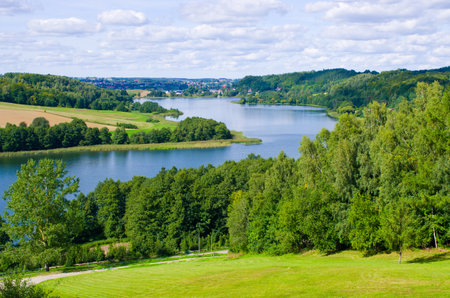 Discover breathtaking summer serenity. Rolling green hills meet a tranquil lake under a vast blue sky, perfect for evoking peace and natural beauty in your projects.の写真素材