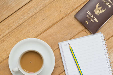 Top view of hot coffee on white cup, passport and pencil on notebook on wood textures is diagonal for backgroundの写真素材