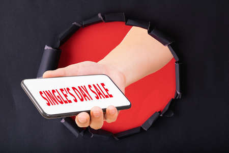 11.11 Single day sale. Hand hold smartphone through red paper torn with Single day sale on screen smart phoneの写真素材