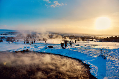 Winter landscape with geothermal springs, tourists, and a setting sun casting a warm glow over the snowy scene. Location: Strokkur Geyser, Iceland.の写真素材