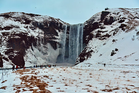 Tourists at a majestic icy waterfall with snow-covered cliffs and a partly cloudy sky. Location: Skogafoss Waterfall Iceland.の写真素材
