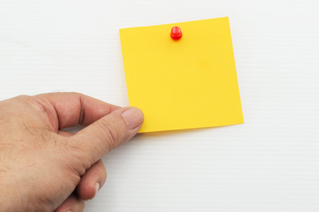 A hand is positioned to hold a bright yellow post note paper fastened with a red pin against a plain white background.の写真素材