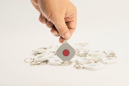 A hand holds an emergency BLE beacon tag above other similar tags resting on a light surface,...
