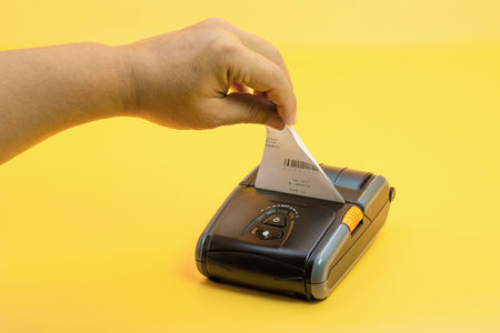 A hand is pulling out a printed receipt from a compact mobile printer placed on a bright yellow...