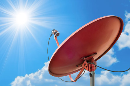 A satellite dish antenna stands prominently against a bright blue sky, illuminated by sunlight and dotted with fluffy clouds.の写真素材
