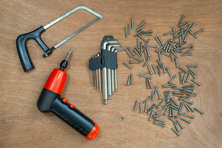 An electric screwdriver sits beside a hacksaw and an assortment of screws on a wooden surface, ready...