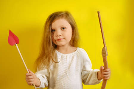 Portrait of happy smiling kid girl holding an arrow in the form of a heart and a crossbow on yellow background with copy space. V2の写真素材