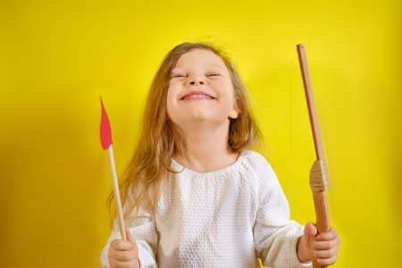 Portrait of happy kid girl holding an arrow in the form of a heart and a crossbow on yellow background with copy space.の写真素材
