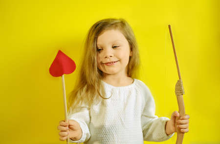 Portrait of kid girl holding an arrow in the form of a heart and a crossbow on yellow background with copy space.の写真素材