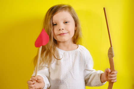 Portrait of smiling kid girl holding an arrow in the form of a heart and a crossbow on yellow background with copy space.の写真素材