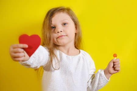 Portrait of smiling kid girl holding a red heart for you and candy in the second hand, isolated on yellow background with copy space. V2の写真素材