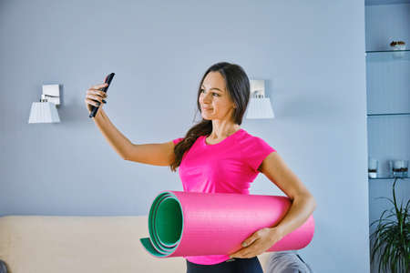 Yoga at home concept. Attractive young woman doing yoga exercises, holds a smartphone and takes a selfieの写真素材