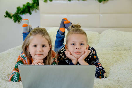 Children lie on the couch in Christmas clothes, and make an online call on a laptop and look in...