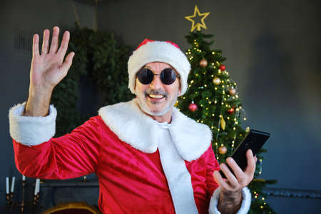 Cheerful Santa Claus in black glasses is sitting in living room and chatting by smartphone with video chat. New year conceptの写真素材