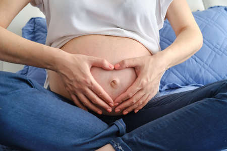 Close up view of young pregnant woman stroking big belly on couch and making heart shape with hands Version 3の写真素材