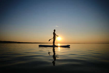 Siluet of Woman standing firmly on inflatable SUP board and paddling through shining water surface. Wide shotの写真素材
