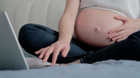 Close up view of Pregnant Woman Resting on a Sofa at Home and Working Remotely with Laptop Computer...