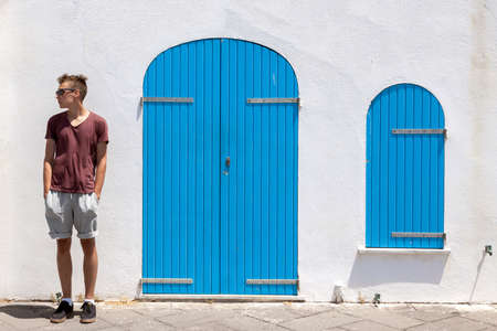 Teenage boy with sunglasses standing relaxed and looking. Mediterranean style white building with closed blue colored doors and window in backgroundの写真素材