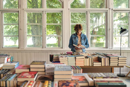 Nida, Lithuania - 25 July, 2020: Stylish middle aged caucasian woman reading a book in old vintage bookshopのeditorial素材