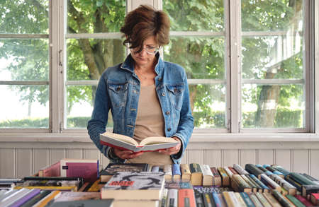 Nida, Lithuania - 25 July, 2020: Closeup on stylish middle aged caucasian woman reading a book in old vintage bookshopのeditorial素材