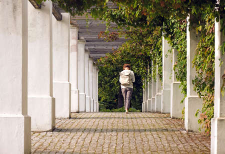 Caucasian woman walking away in outdoor alley overgrown with plants in autumnの写真素材