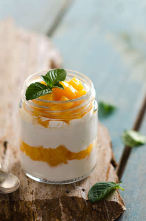 Plain yoghurt with pieces of peach and mint leaves in a jar on a yellow stone and light blue wooden backdrop.の写真素材