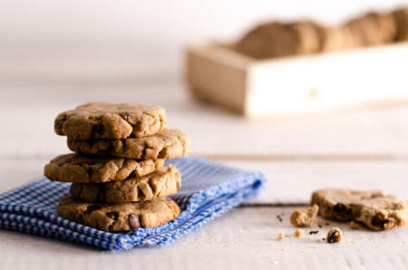 Vanilla and chocolate cookies stacked on a white and blue napkin, a bitten cookie and cookies in a wooden box, on a white wooden backdrop.の写真素材