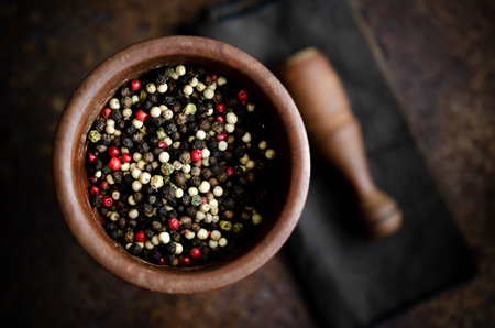 Black, white, green, and pink peppercorns in a wooden mortar with pestle on a brown napkin and dark backdrop.の写真素材