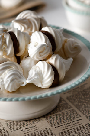 White meringue cookies with dulce de leche on a white and green cake stand on an old magazine and some tea cups in the background.の写真素材