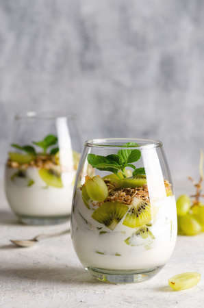 Two glasses of plain yoghurt with pieces of fresh kiwi fruits, white grapes, granola, and mint leaves on a gray background.の写真素材
